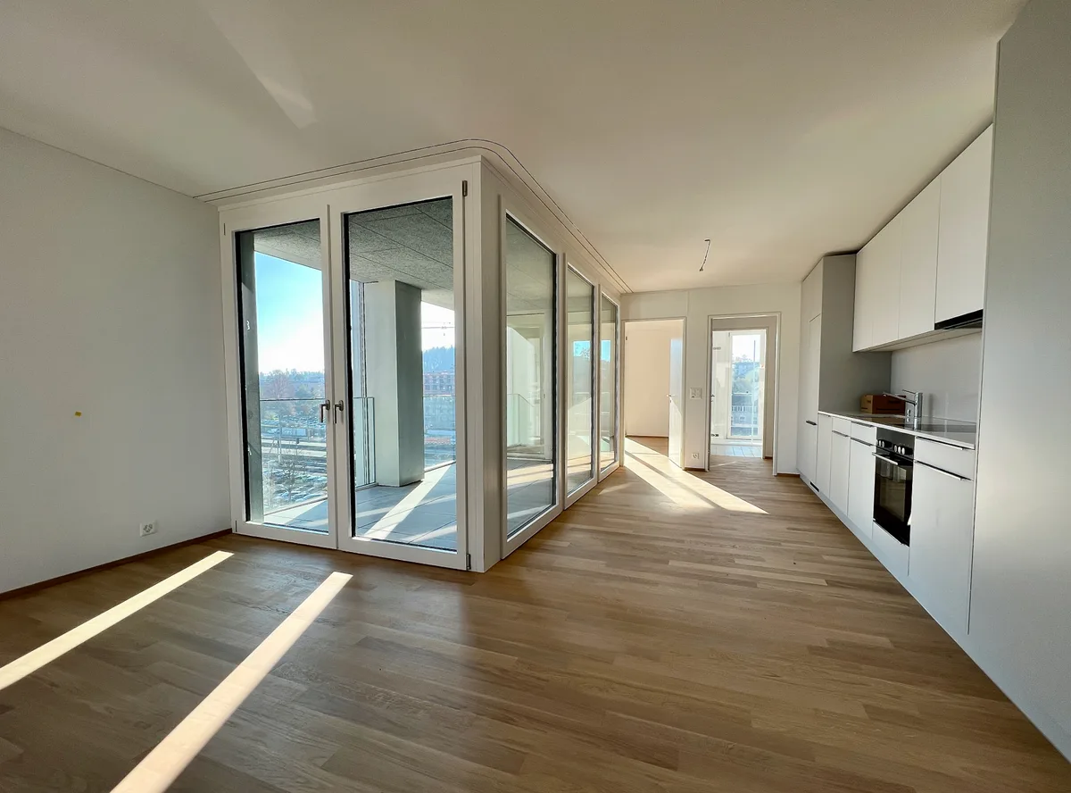 Man sieht den Wohn- und Küchenraum einer Wohnung. Gleich gegenüber der Küchenzeile geben bodenlange Fenster und Türen den Blick auf den Balkon frei. Im Hintergrund stehen die Türen zu einem weiteren Zimmer und zum Badezimmer offen.